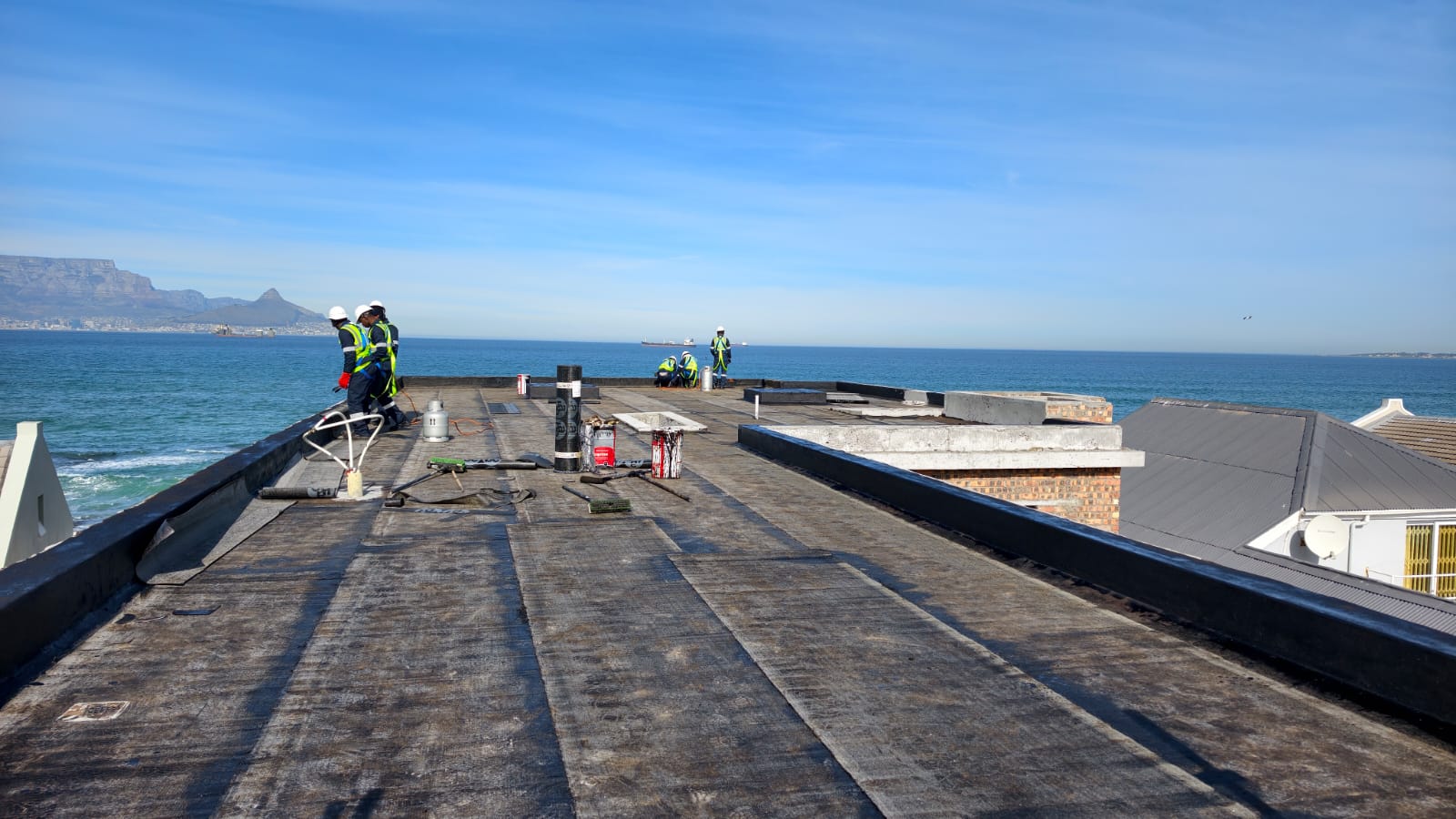 Long flat roof stripped back to the old bitumen, crew starting at the seaward parapet