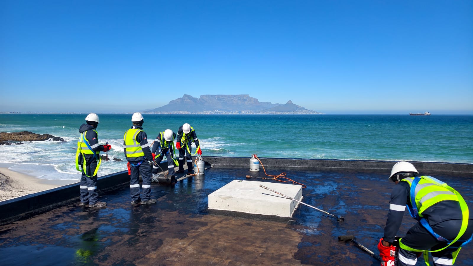 Crew in harnesses and hard hats bonding torch-on membrane along a beachfront flat roof, Table Mountain across the bay