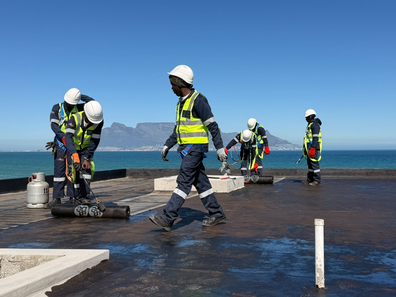Membrane roll being bedded down as the crew works across a flat roof, Table Mountain behind them